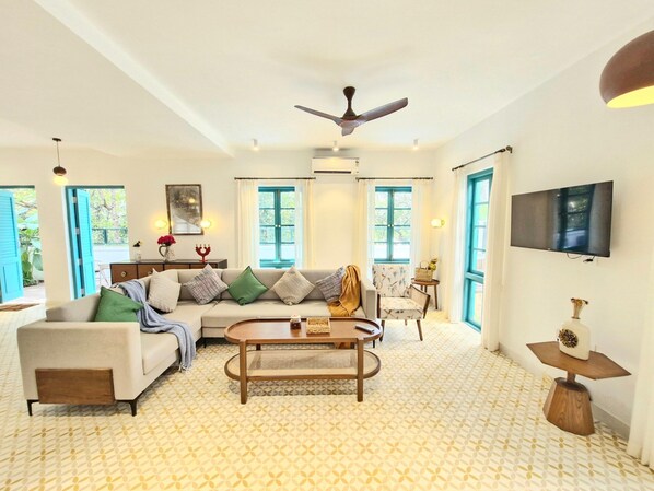 A spacious living room with a sofa, armchair, and coffee table, large windows with blue shutters, and a ceiling fan.