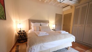 A neatly made bed with white linens and pillows, two bedside tables with lamps, a large wardrobe, and a door to another room.