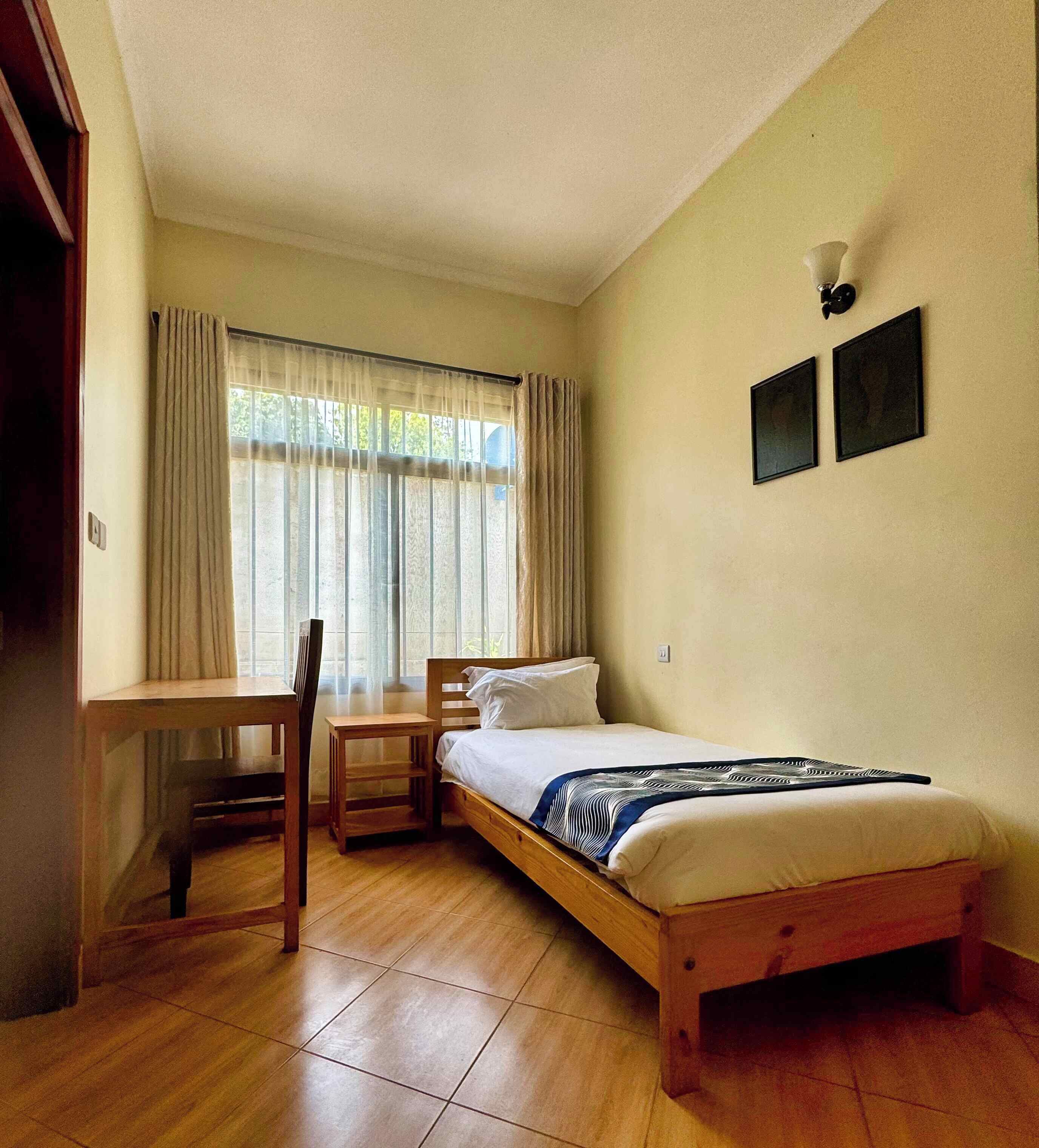 A small, tidy room with a bed, a desk, and a chair. There are two framed pictures on the wall.