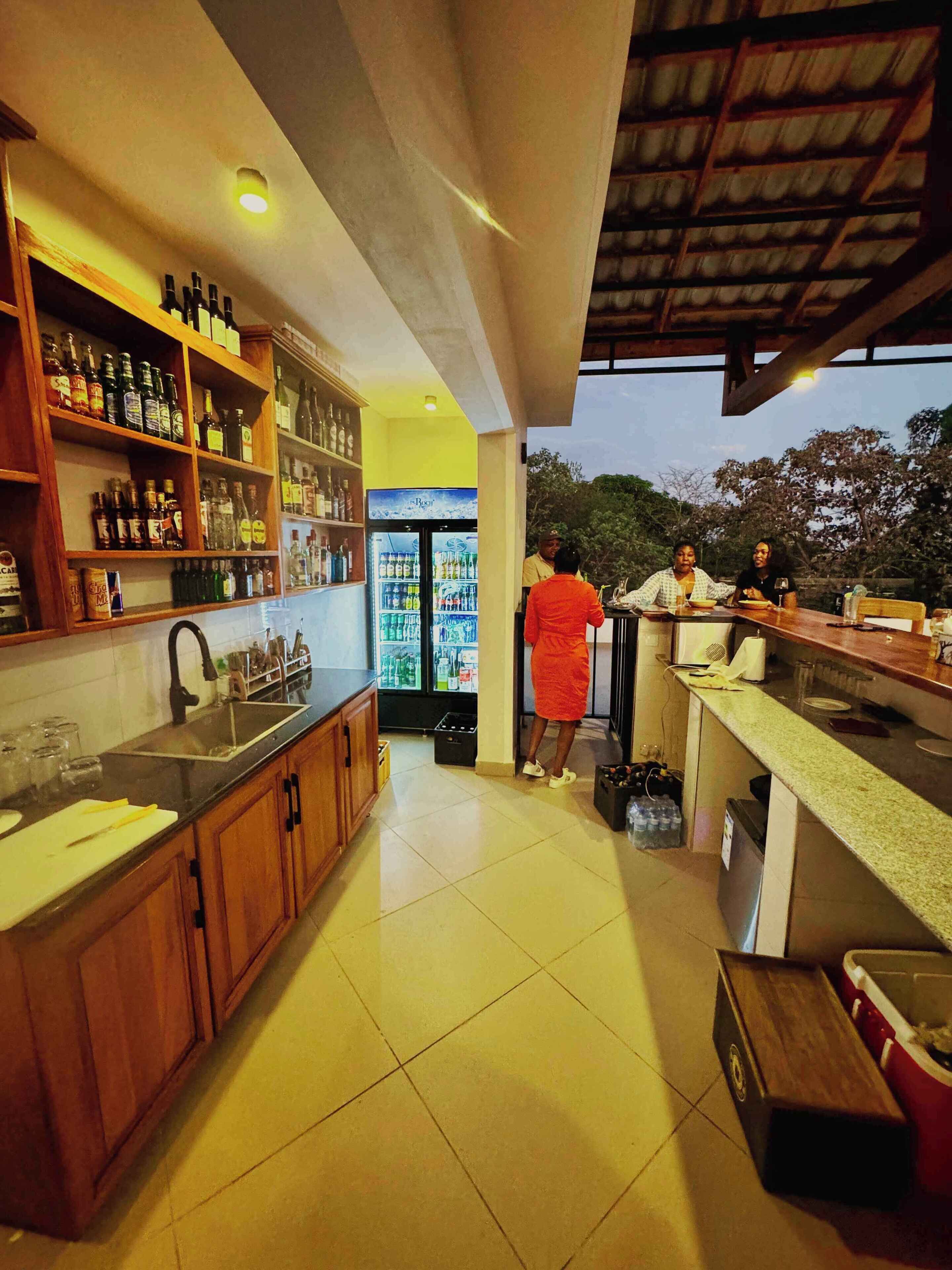 A bar area with shelves of bottles, a refrigerator, and a counter where two people are seated.