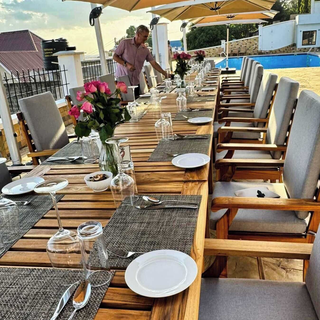 A long wooden dining table set with plates, cutlery, and glassware, surrounded by cushioned chairs, with a pool and a building in the background.