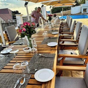 A long wooden dining table set with plates, cutlery, and glassware, surrounded by cushioned chairs, with a pool and a building in the background. - Moshono Hillside  (Arusha)