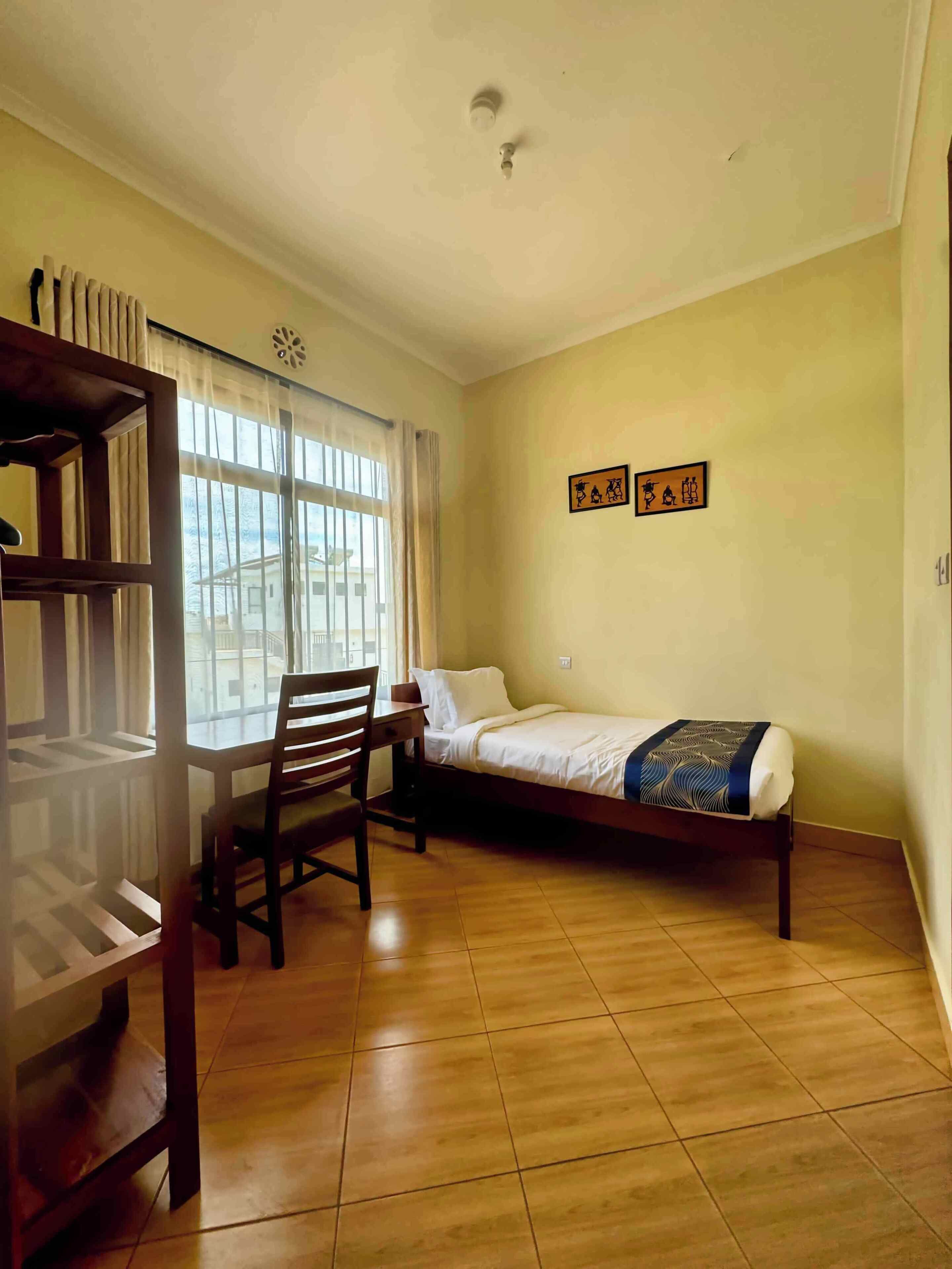 A room with a bed, a desk, and a chair. There are two windows with curtains, and a wall with two framed pictures.