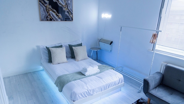Une chambre moderne avec un grand lit, un radiateur mobile, un canapé gris et une fenêtre avec des stores.