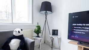 Un salon moderne avec un canapé, un ours en peluche à l’effigie d’un panda, un petit arbre, une lampe sur pied et un téléviseur qui affiche un message.