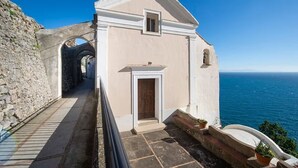 Un estrecho sendero de piedra conduce a un pequeño edificio de fachada blanca y una ventana, con vistas al mar azul y cristalino.