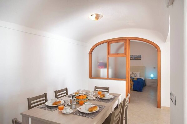 Un coin repas avec une table mise pour le petit-déjeuner, un lit visible à travers une porte en arc de cercle, et une table de chevet avec une lampe.