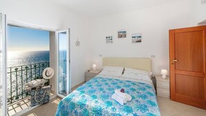 Un dormitorio con cama, mesita de noche, puerta de madera y vistas al mar desde un balcón.