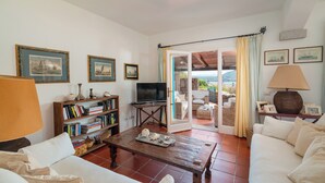 A living room with a television, bookshelves, a coffee table, and a view through a glass door to an outdoor patio. - Casa Turchese by Wonderful Italy (Olbia)