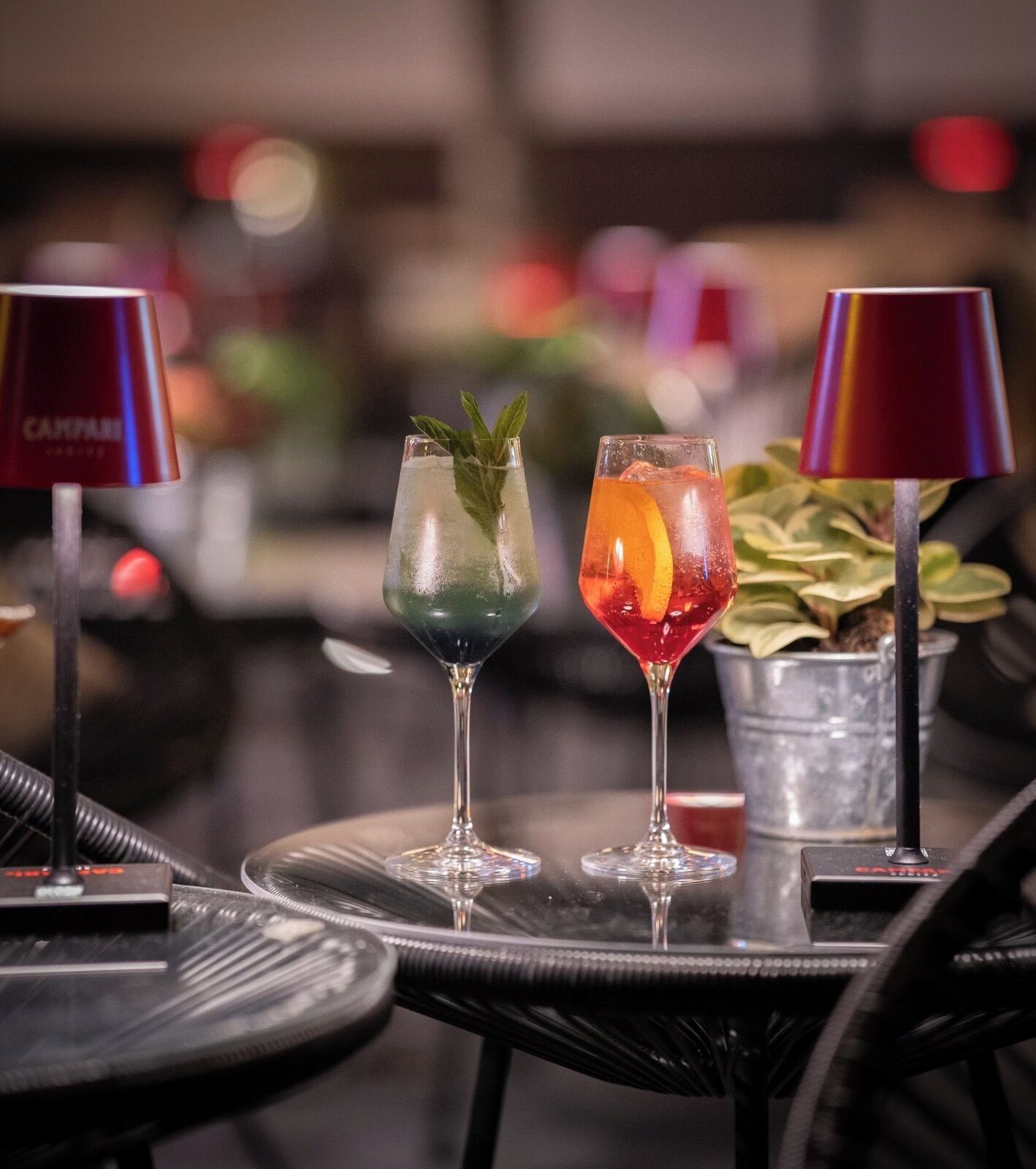 Two cocktails on a table with a metal bucket and a potted plant in the background.