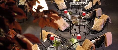 Een terras met ronde tafels en zwarte draadstoelen, omgeven door herfstkleurige bladeren.