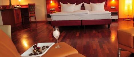 A hotel room with a large bed, a bedside table with a lamp, a small table with a plate of chocolates, and a glass of dessert.