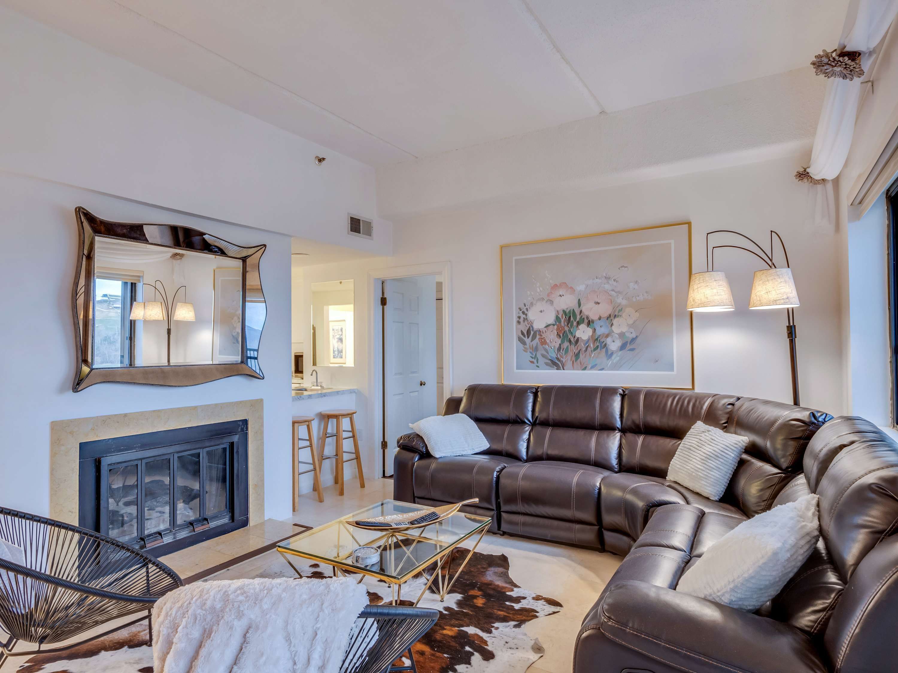 A modern living room with a leather sofa, a glass coffee table, a fireplace, and a large mirror.