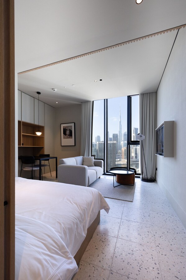 Ein modernes Hotelzimmer mit einem großen Bett, einer Sitzecke mit Couchtisch, einem Schreibtisch mit Stühlen, einem Fernseher und Blick auf die Stadt durch große Fenster.