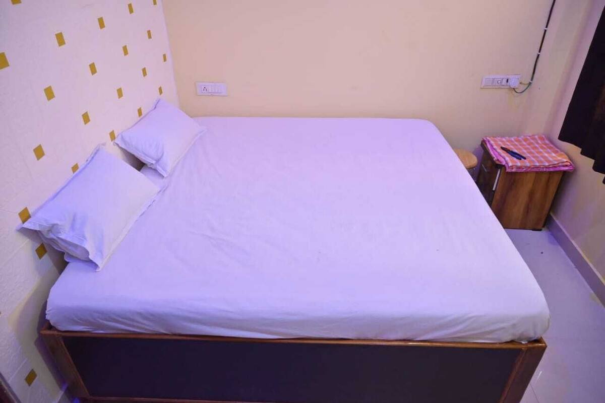 Ein Einzelbett mit weißer Matratze und Kissen, ein hölzernes Kopfende und ein kleiner Nachttisch mit kariertem Stoff in Rosa und Weiß.