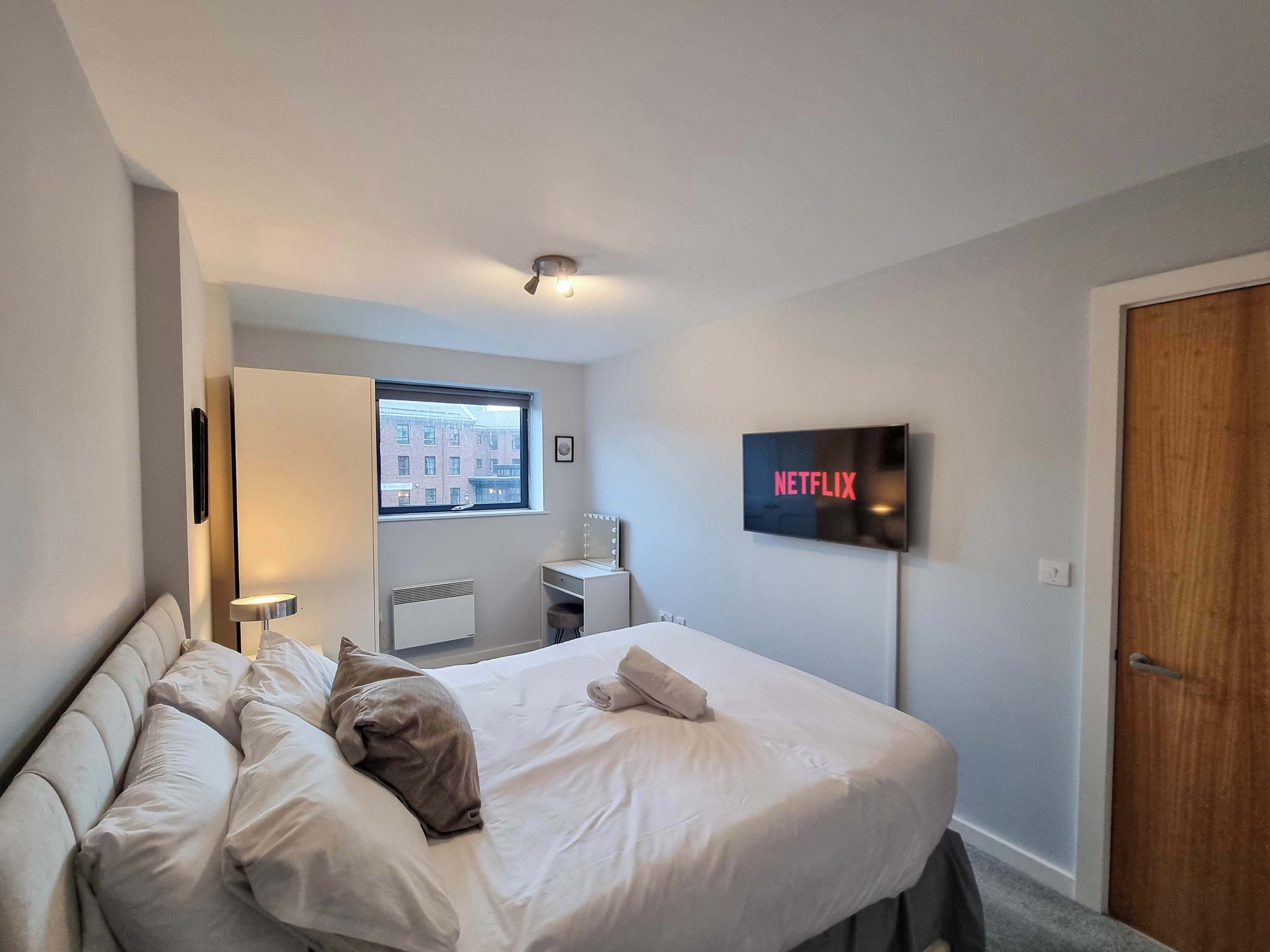 Una habitación de hotel con cama, televisor montado en la pared, ventana con vistas a edificios y puerta.