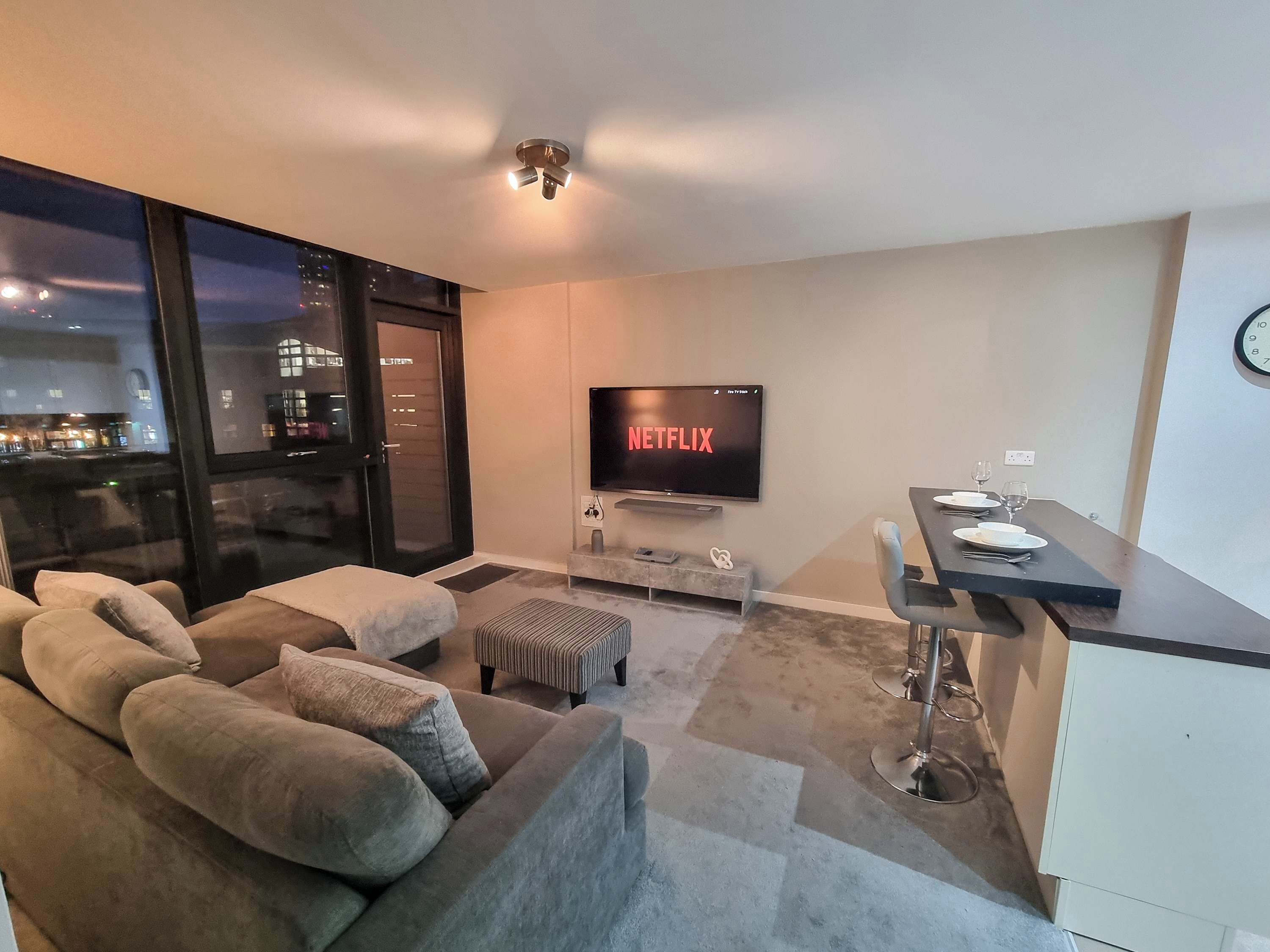Una sala moderna con sofá, televisión mostrando Netflix, comedor con mesa y sillas, y vista a la ciudad por la ventana.