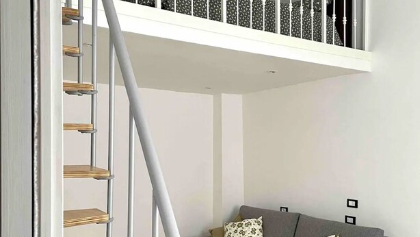 A modern living room with a grey sofa, two patterned cushions, and a wooden staircase with a white railing.
