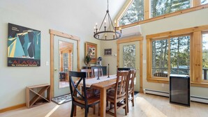A dining area with a wooden table and chairs, a chandelier, and large windows offering a view of trees. - Family Retreat | 10 Min to Okemo (Ludlow)