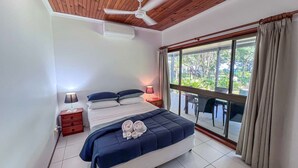 A bedroom with a bed, bedside tables, a ceiling fan, a window with curtains, and a view of greenery outside. - Bluefin House beachfront access (Mission Beach)