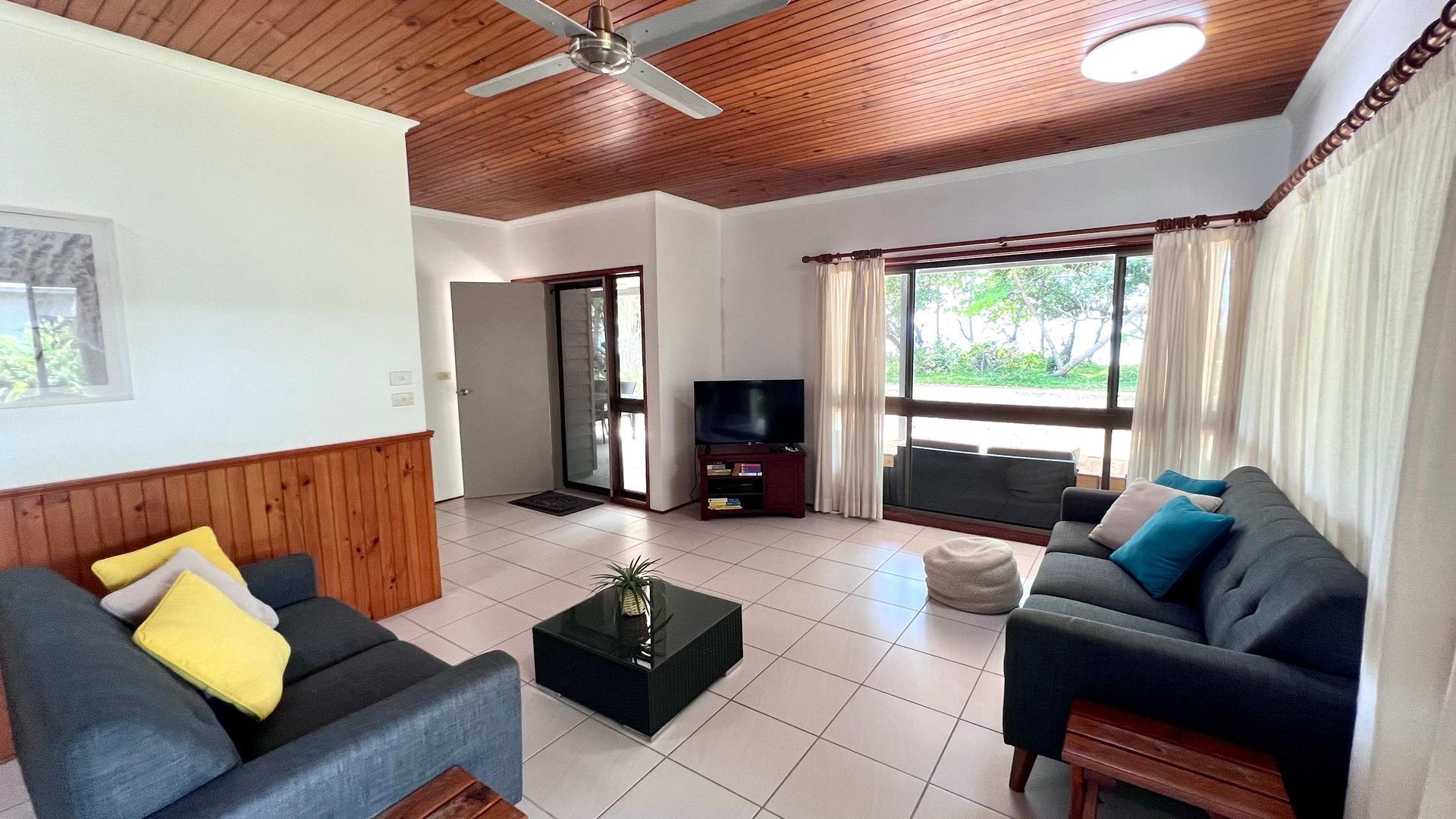 Una sala de estar con ventilador de techo, televisor sobre un mueble bajo, dos sofás con cojines, una mesa de centro y un ventanal con cortinas.