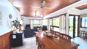 A spacious living area with a wooden ceiling, a dining table with chairs, a sofa, a TV, and large windows. - Bluefin House beachfront access (Mission Beach)