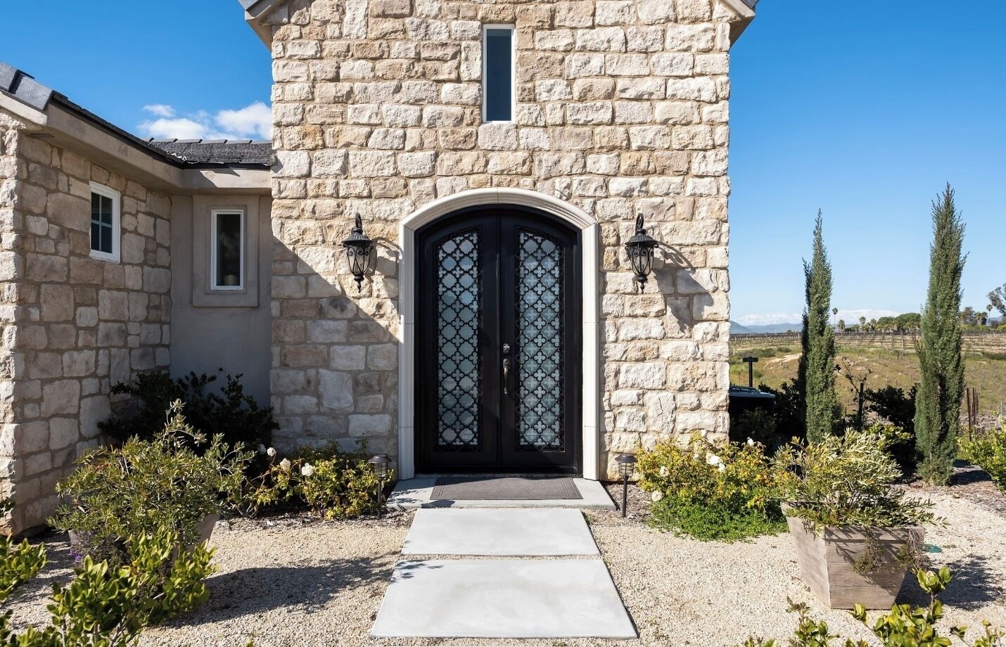 Un edificio de piedra con una puerta doble negra, una ventana pequeña y un camino de grava que conduce a la entrada.