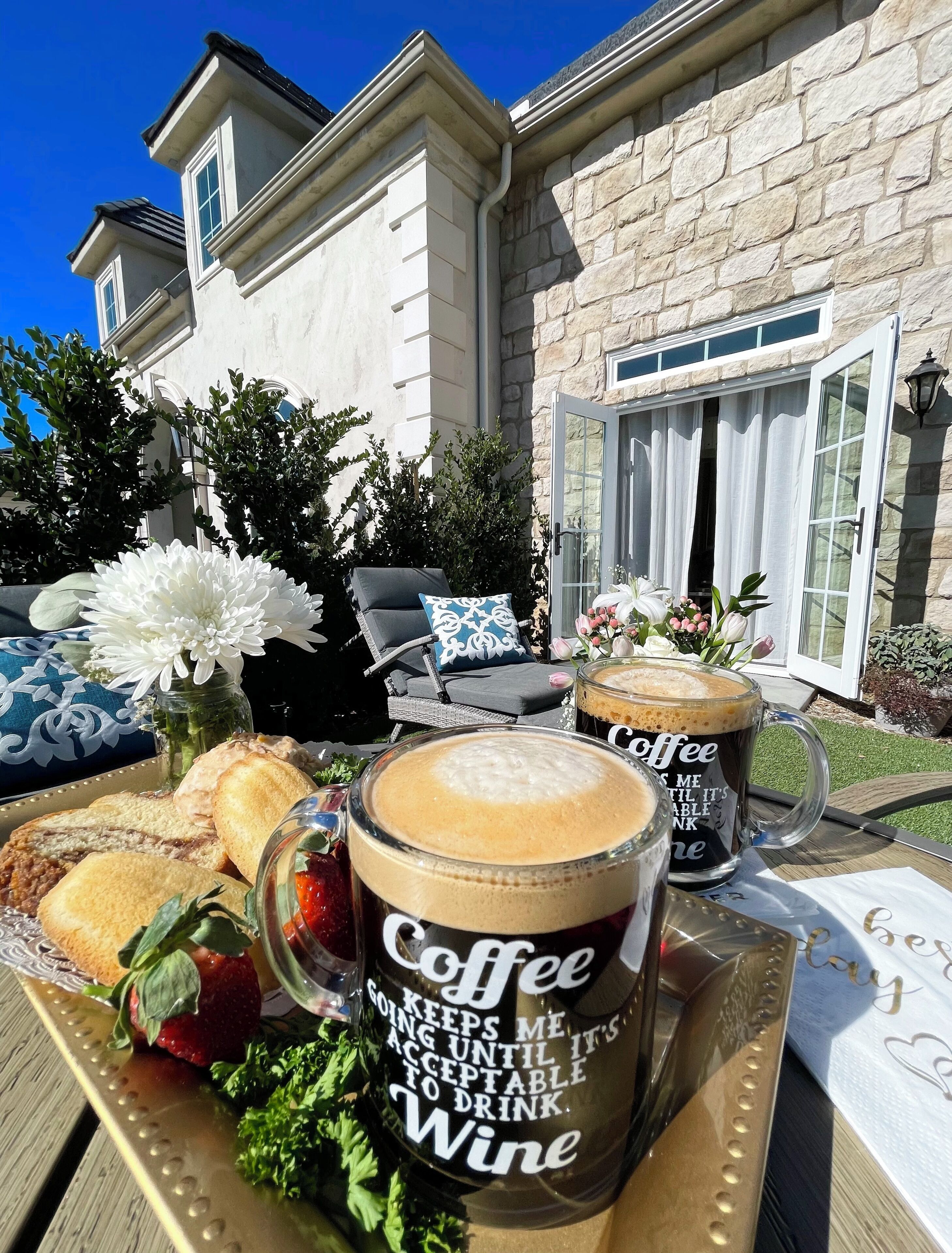 A patio setting with a table set for two, featuring a tray with pastries, a glass of coffee, and a mug with a humorous message.