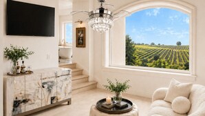 A living room with a marble cabinet, a television, a chandelier, a round ottoman, a sofa, and a large window with a vineyard view. - Domaine Chardonnay (Temecula)