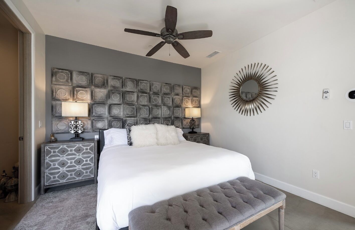 A bedroom with a large bed, a ceiling fan, a decorative wall panel, a round mirror, and a bench.