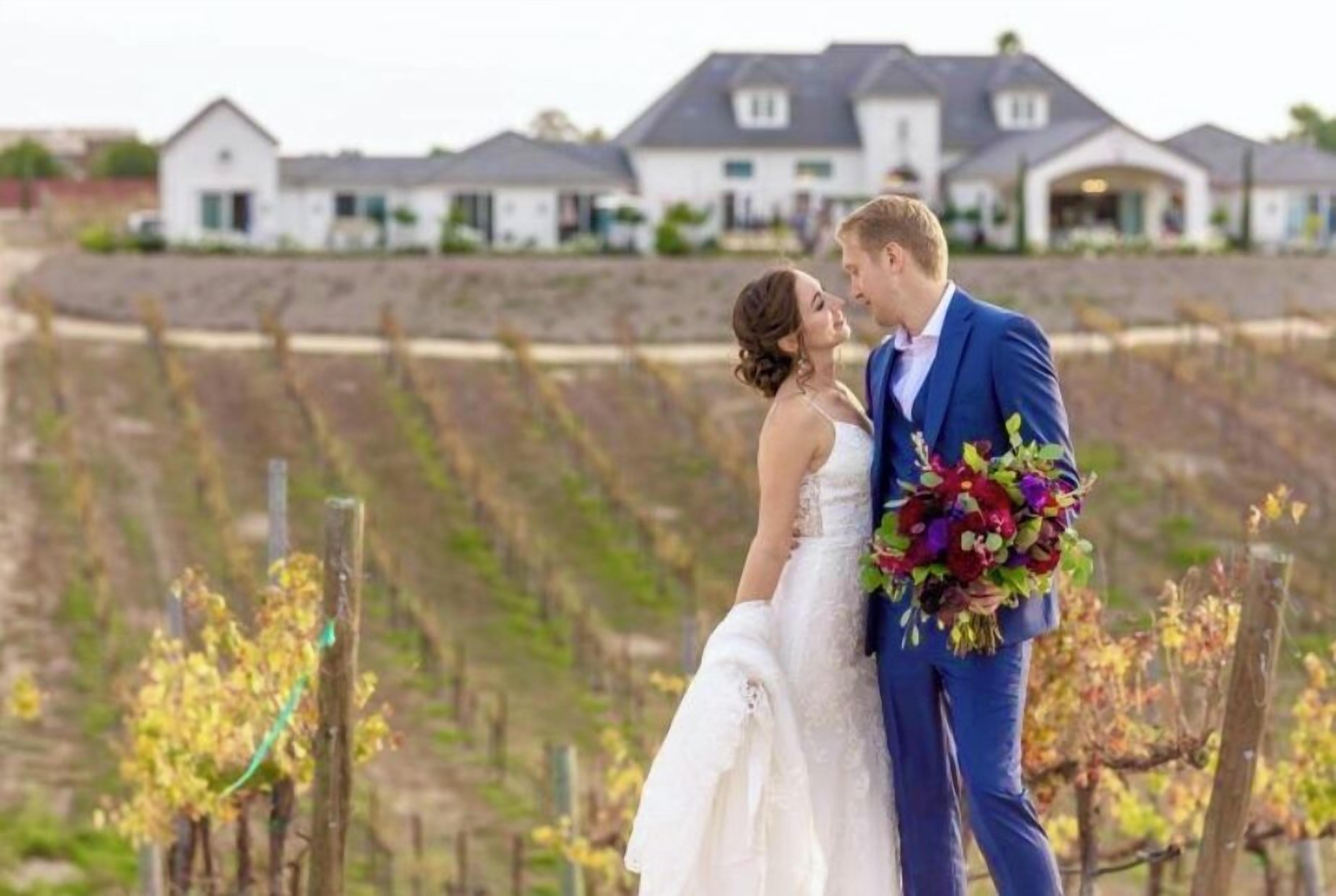 A couple embracing in a vineyard setting with houses in the background.