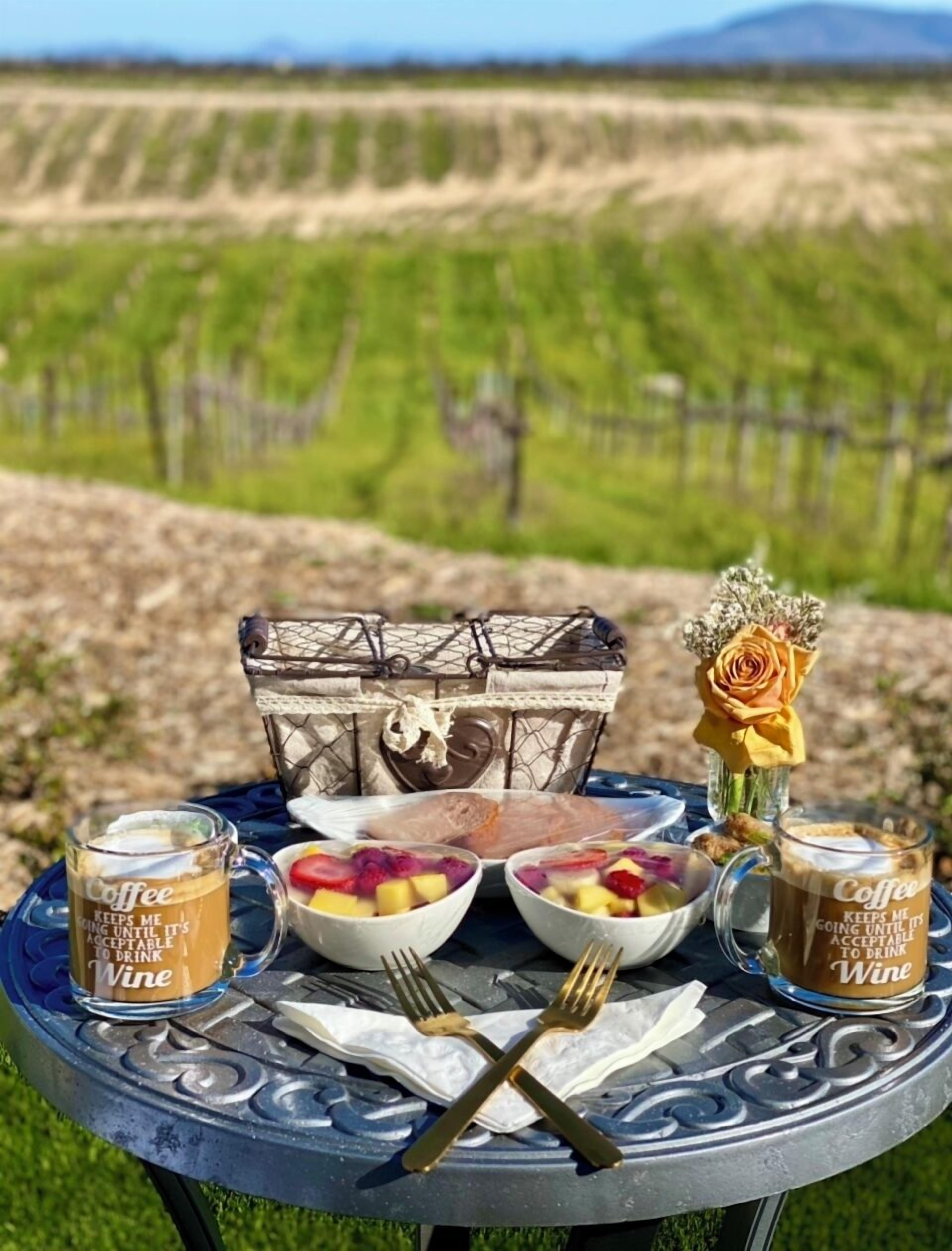 A table set outdoors with a view of a vineyard, a basket, a vase with a rose, and two coffee mugs.