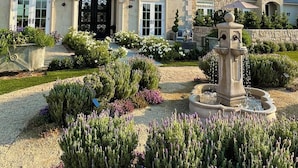 A stately house with a prominent black front door, a well-maintained garden with a fountain, and a hot air balloon in the sky. - Domaine Chardonnay (Temecula)