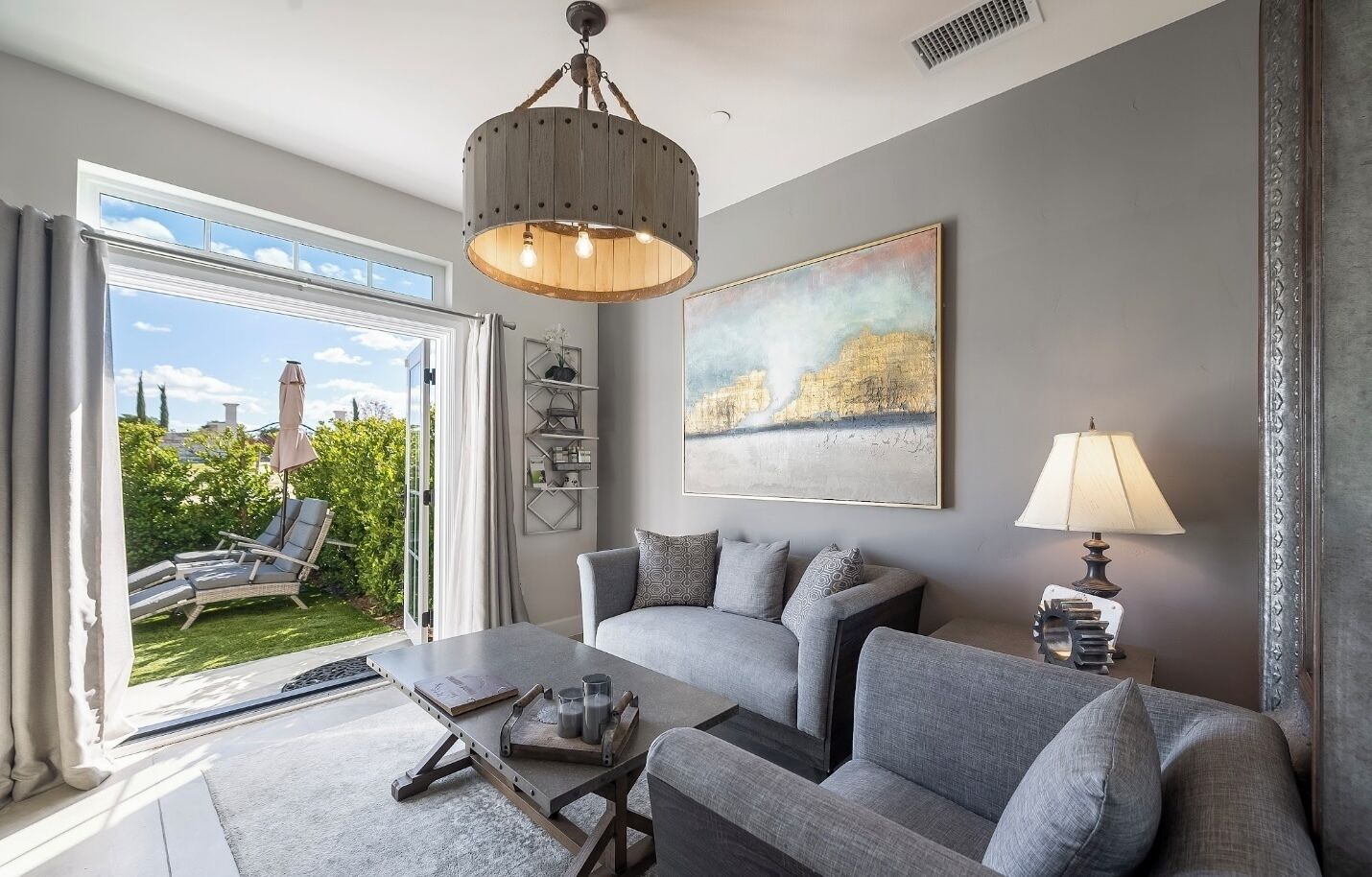 A modern living room with a grey sofa set, a coffee table, and a chandelier. There is a large window with a view of greenery and a building outside.