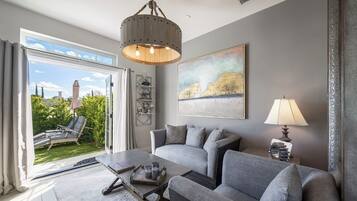 A modern living room with a grey sofa set, a coffee table, and a chandelier. There is a large window with a view of greenery and a building outside.