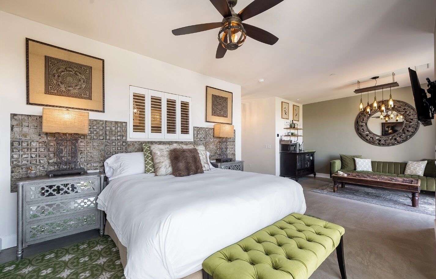 A spacious bedroom with a large bed, a green bench, a ceiling fan, and a dining area with a chandelier.