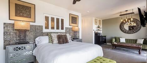 A spacious bedroom with a large bed, a green bench, a ceiling fan, and a dining area with a chandelier.