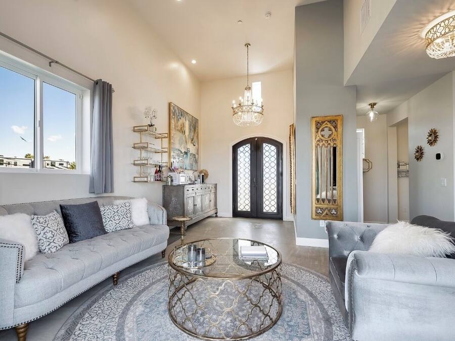 A spacious living room with a grey sofa, a round coffee table, and a grey armchair. There is a large window with curtains, a chandelier, and a decorative mirror.