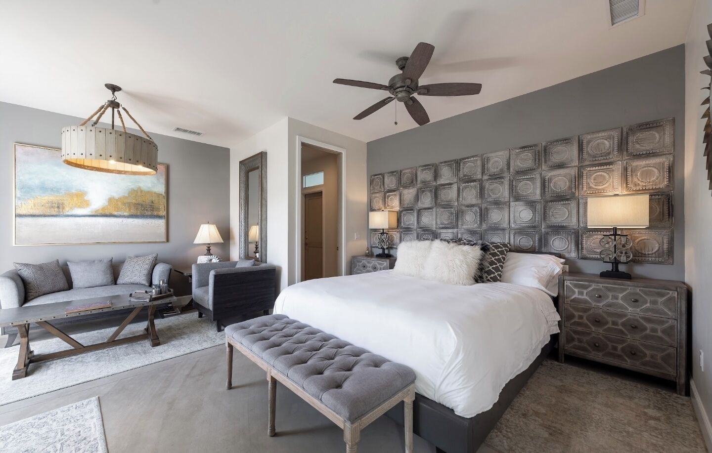 Een moderne slaapkamer met een groot bed, een plafondventilator, een decoratief wandpaneel en een zithoek met een bank en een bijzettafeltje.