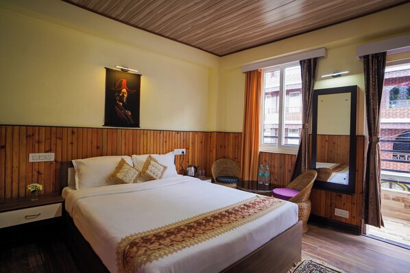 A hotel room with a large bed, wooden paneling, a wall-mounted painting, a window with curtains, and a small table with a chair. - Hotel Hill Queen (Darjeeling)