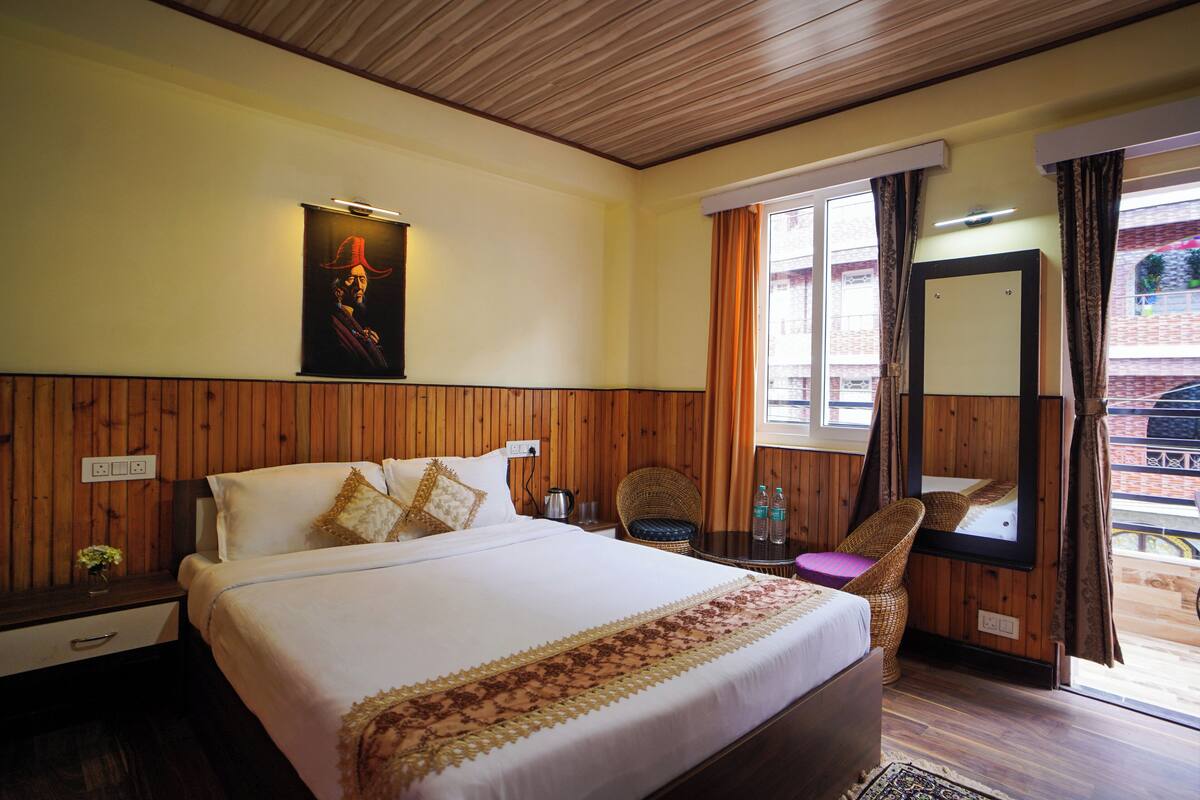 Een hotelkamer met een groot bed, een houten hoofdeinde, een tafeltje met een stoel, een raam met gordijnen, en een spiegel.