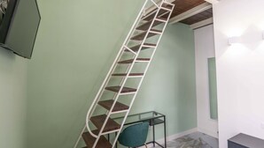 Un escalier en colimaçon avec une structure en métal blanc et des marches en bois, menant à un palier où se trouvent une table en verre et une chaise de couleur turquoise.