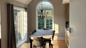 Une salle à manger spacieuse avec un haut plafond et une grande fenêtre, une cuisine moderne dotée d’un plan de travail en bois, et un canapé en cuir dans un salon lumineux.