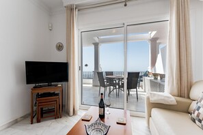 Un salon moderne doté d’un téléviseur à écran plat, de meubles en bois, d’une porte vitrée donnant sur un balcon avec coin salon extérieur et vue sur la mer.