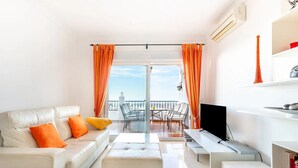 Un salon moderne avec un canapé, un pouf et un meuble pour la télévision. Il y a un balcon avec une table et des chaises.