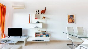 Un salon moderne avec un téléviseur à écran plat, des étagères blanches ornées d’objets décoratifs, une table à manger en verre et un canapé blanc.