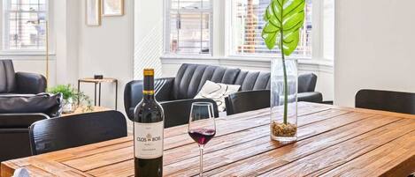 A wooden dining table with a bottle of Clos du Bois wine and a glass of red wine, a potted plant, and a modern living room in the background.
