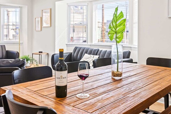 A wooden dining table with a bottle of Clos du Bois wine and a glass of red wine, a potted plant, and a modern living room in the background.