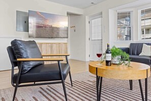 Un salon moderne avec un fauteuil en cuir, une table basse ronde en bois sur laquelle se trouvent une bouteille de vin et un verre, un téléviseur à écran plat fixé au mur et un canapé.