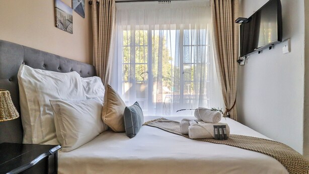 Una cama bien tendida con almohadas, una mesita de noche con lámpara, un televisor montado en la pared y una ventana con cortinas.